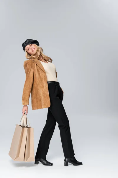 Full length of blonde woman in stylish leather beret and beige blazer posing with shopping bags on grey — Fotografia de Stock