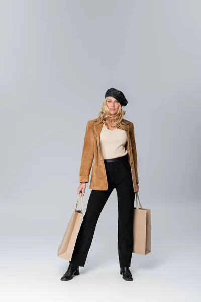 Full length of happy woman in stylish leather beret and beige blazer holding shopping bags on grey — Stock Photo