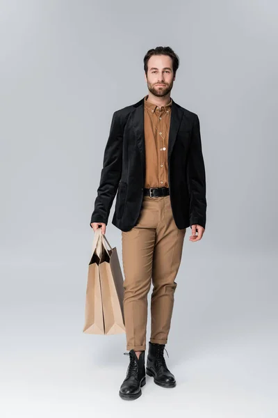 Full length of bearded man in stylish autumnal outfit posing with shopping bags on grey — Stockfoto