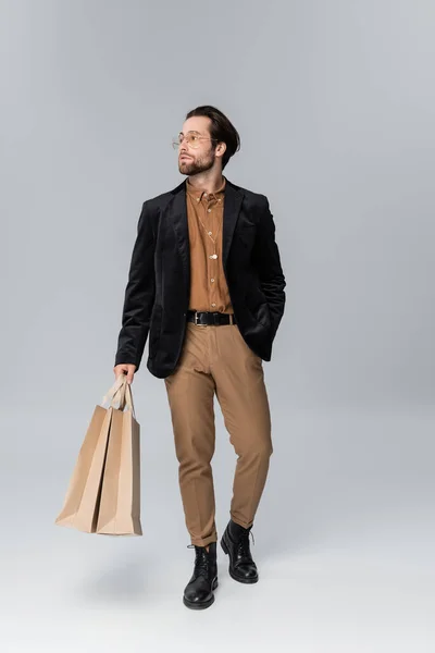 Full length of bearded man in sunglasses and stylish autumnal outfit posing with shopping bags on grey - foto de stock