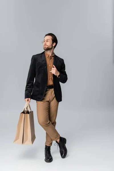 Full length of bearded man in sunglasses and stylish autumnal outfit posing with paper bags on grey — Stockfoto