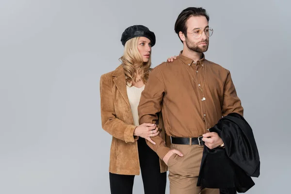 Blonde woman in stylish beret hugging bearded man in sunglasses and posing with hand in pocket isolated on grey — Stockfoto