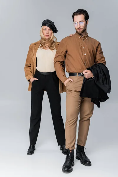 Full length of blonde woman in stylish beret hugging bearded man in sunglasses and posing on grey — Stockfoto