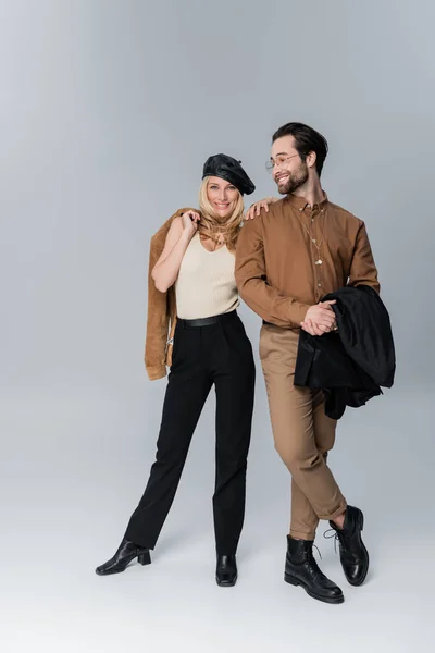 Full length of happy woman in stylish beret hugging bearded man in sunglasses and posing on grey — Stock Photo