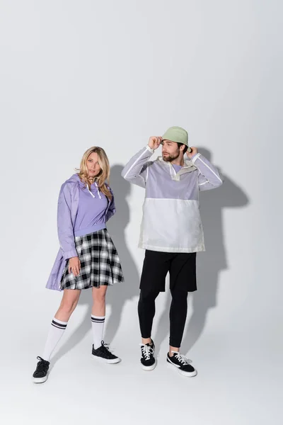 Full length of stylish woman in tartan skirt and longs socks with sneakers posing near man in panama hat on grey — Fotografia de Stock