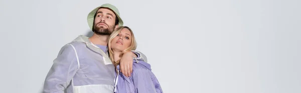 Bearded man in panama hat hugging blonde girlfriend in purple raincoat isolated on grey, banner — Stockfoto