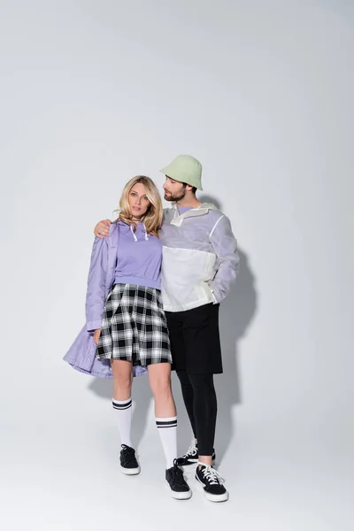 Full length of bearded man in panama hat hugging stylish woman in tartan skirt and longs socks with sneakers on grey — Fotografia de Stock