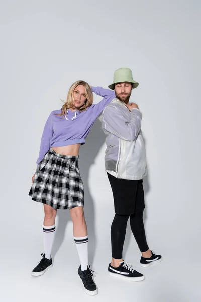 Full length of stylish woman in tartan skirt and longs socks with sneakers leaning on near man in panama hat on grey — Stockfoto