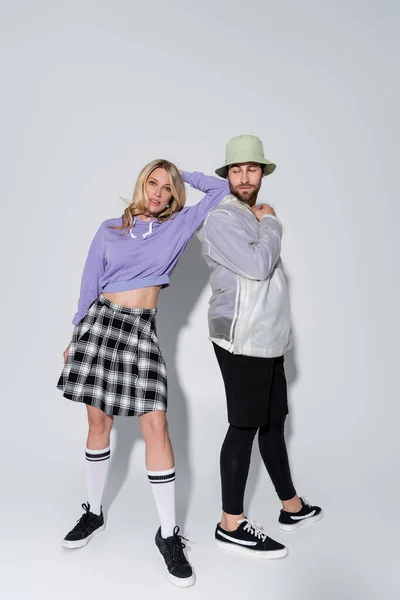 Full length of stylish woman in tartan skirt and longs socks with sneakers leaning on near man in panama hat on grey — Foto stock