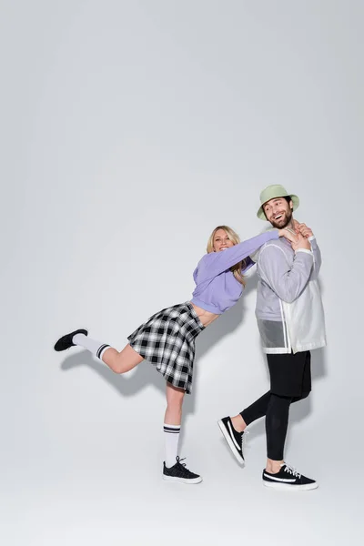Full length of happy woman in tartan skirt and longs socks with sneakers leaning on man in panama hat on grey — Stockfoto