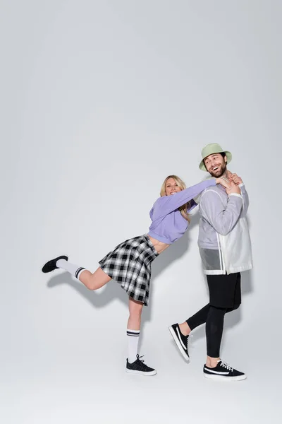 Full length of happy woman in tartan skirt and longs socks with sneakers leaning on man in panama hat on grey — Photo de stock