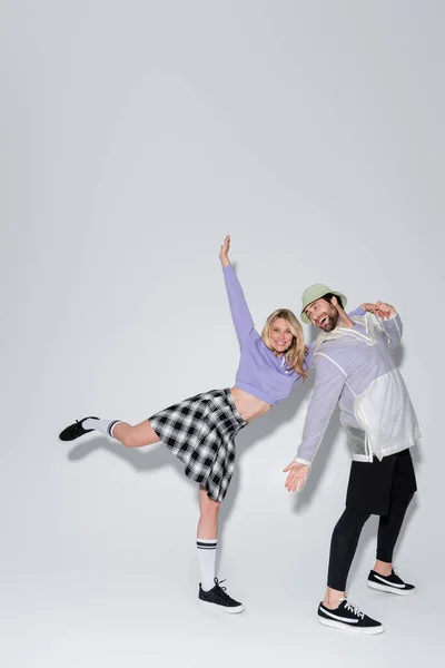 Full length of cheerful woman in tartan skirt and longs socks with sneakers leaning on man in panama hat on grey - foto de stock