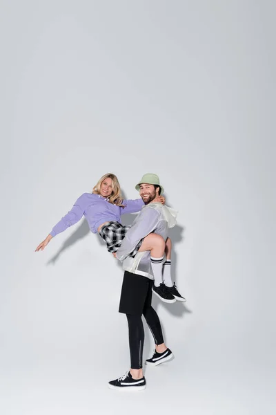 Full length of happy man in panama hat holding in arms woman in tartan skirt and longs socks on grey — Stock Photo