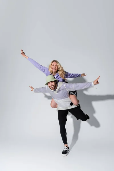 Full length of cheerful woman and happy man in panama hat posing with outstretched hands on grey — Fotografia de Stock