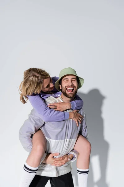 Happy man in panama hat sticking out tongue and piggybacking woman in longs socks on grey - foto de stock
