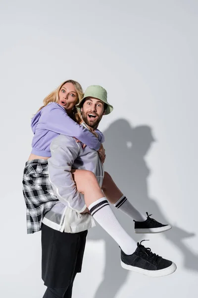 Amazed man in panama hat piggybacking surprised woman in longs socks and sneakers on grey — Stock Photo