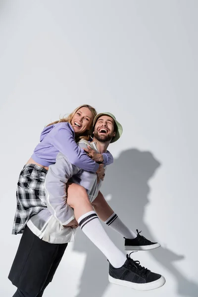 Amazed man in panama hat piggybacking joyful woman in longs socks and sneakers on grey — Photo de stock