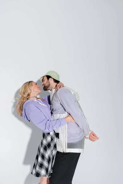 Bearded man in panama hat hugging happy blonde girlfriend in tartan skirt on grey - foto de stock