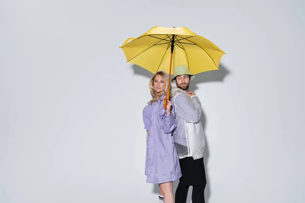 Blonde woman in tartan skirt standing with bearded man in panama hat under yellow umbrella on grey - foto de stock