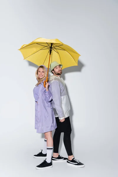 Full length of happy woman in tartan skirt standing with bearded man in panama hat under yellow umbrella on grey - foto de stock