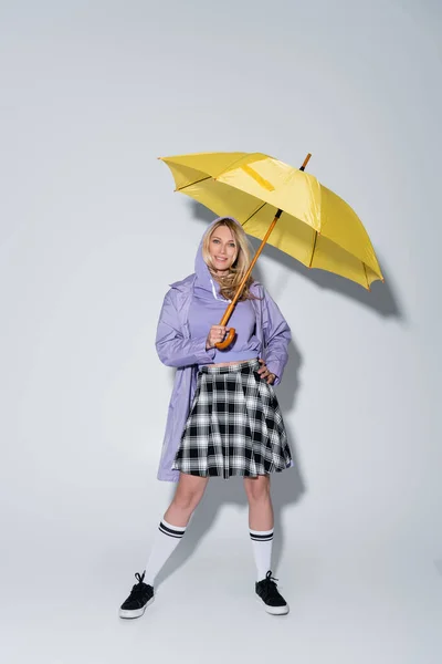 Full length of happy woman in tartan skirt and longs socks with sneakers standing under umbrella on grey — Stock Photo