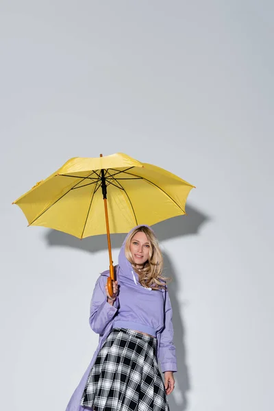 Happy woman in tartan skirt smiling standing under yellow umbrella on grey — Stockfoto