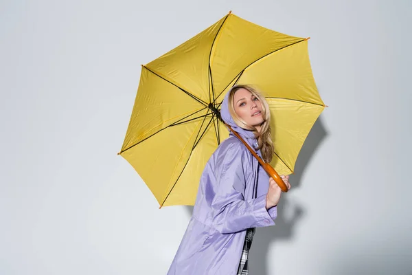 Blonde woman purple rain jacket standing under yellow umbrella on grey — Stock Photo