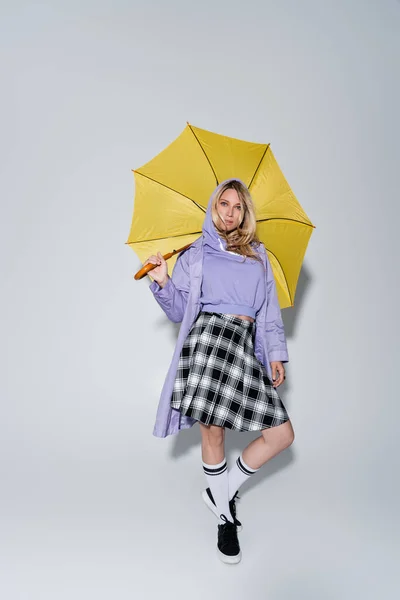 Full length of blonde model in tartan skirt and longs socks with sneakers standing under yellow umbrella on grey — Photo de stock