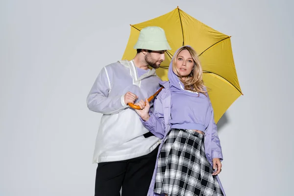 Woman in tartan skirt posing with man in panama hat under yellow umbrella on grey — Stock Photo