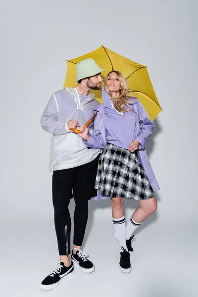 Full length of woman in tartan skirt and longs socks posing with man in panama hat under yellow umbrella on grey — Photo de stock