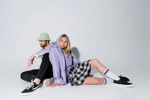 Full length of blonde woman in tartan skirt and longs socks sitting with stylish man in panama hat on grey — Stock Photo