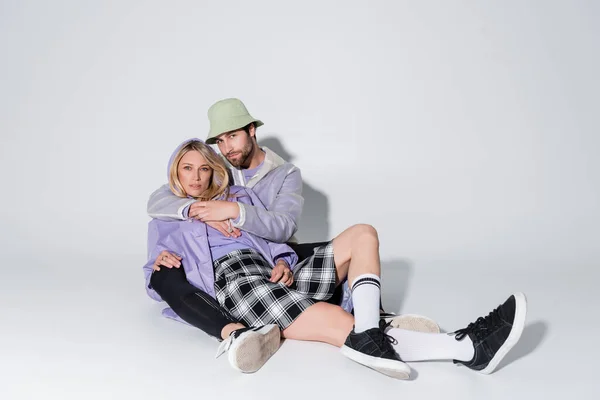 Full length of stylish man in panama hat hugging blonde woman in tartan skirt and longs socks while sitting on grey - foto de stock