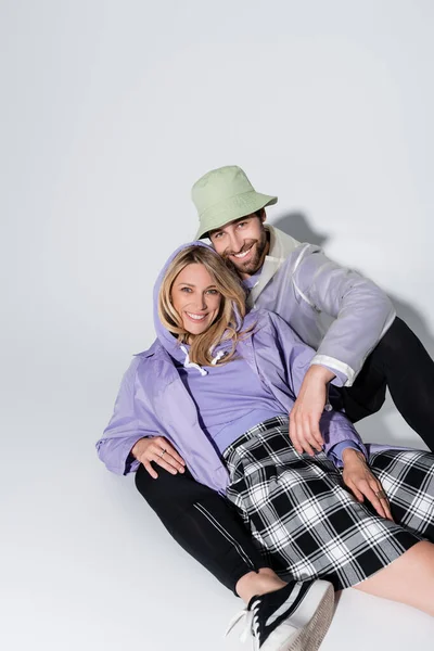 Cheerful man in panama hat hugging blonde woman in tartan skirt while sitting on grey — Photo de stock