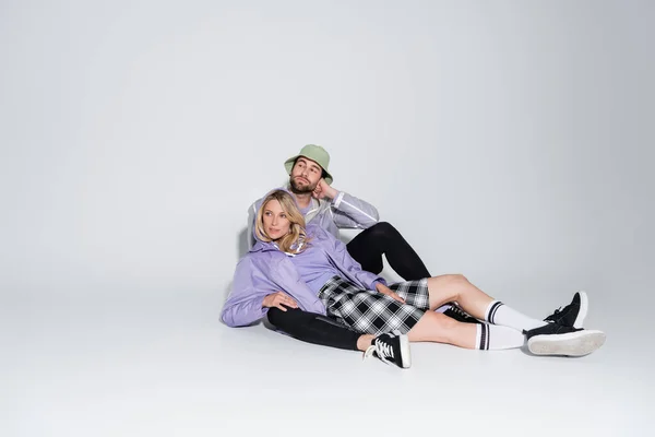 Full length of man in stylish panama hat hugging blonde woman in tartan skirt and longs socks while sitting on grey — Photo de stock