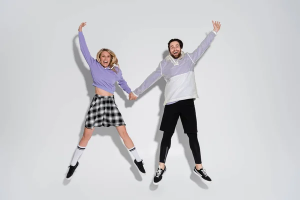 Full length of excited man in leggings and shorts jumping with blonde woman in tartan skirt on grey — Stockfoto