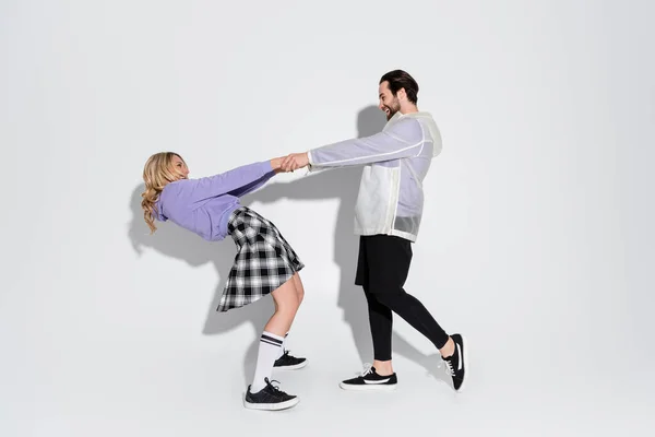 Full length of happy man and blonde woman in tartan skirt holding hands on grey - foto de stock