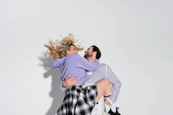 Cheerful man holding in arms smiling blonde woman in tartan skirt on grey - foto de stock