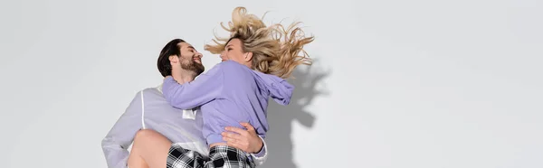 Cheerful man holding in arms smiling blonde woman in tartan skirt on grey, banner - foto de stock