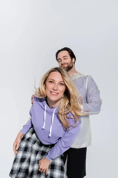 Веселая женщина в фиолетовой толстовке, смотрящая в камеру рядом с мужчиной, изолированным на сером — стоковое фото