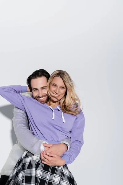 Happy man hugging cheerful woman in purple sweatshirt while looking at camera on grey — Fotografia de Stock