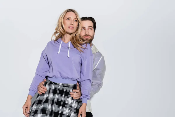 Bearded man hugging cheerful woman in purple sweatshirt while looking away isolated on grey — Stock Photo