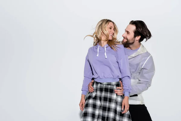 Happy man hugging cheerful woman in purple sweatshirt and skirt while looking at each other on grey — Fotografia de Stock