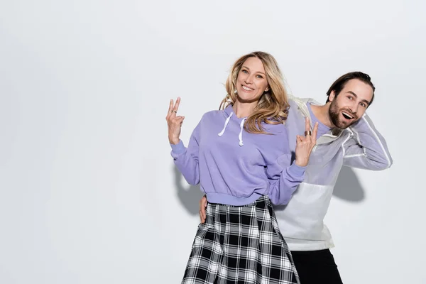 Excited man near cheerful woman in purple sweatshirt showing peace sign on grey — Stockfoto