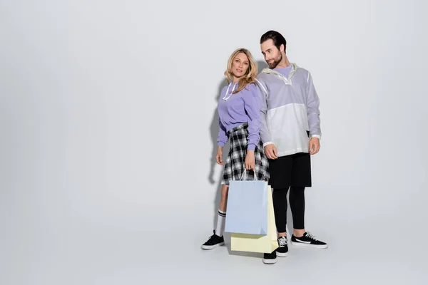 Full length of happy woman in plaid skirt holding shopping bags near bearded man on grey - foto de stock