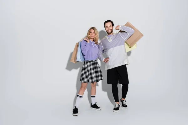 Full length of joyful man and cheerful woman in tartan skirt holding shopping bags on grey — Foto stock