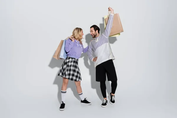 Full length of excited man and happy woman in tartan skirt holding shopping bags on grey — Stockfoto