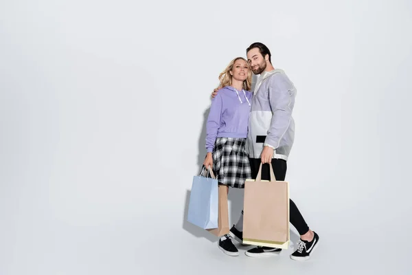 Full length of happy man and blonde woman in tartan skirt holding shopping bags on grey — Stock Photo