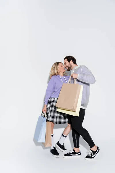Full length of happy man holding shopping bags near blonde woman in tartan skirt on grey - foto de stock