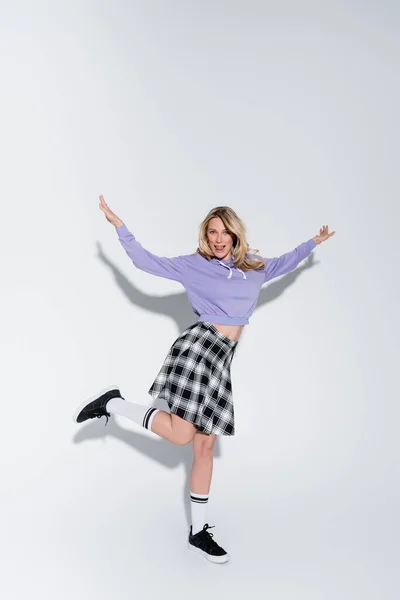 Full length of cheerful blonde woman in trendy outfit posing with outstretched hands on grey — Stock Photo