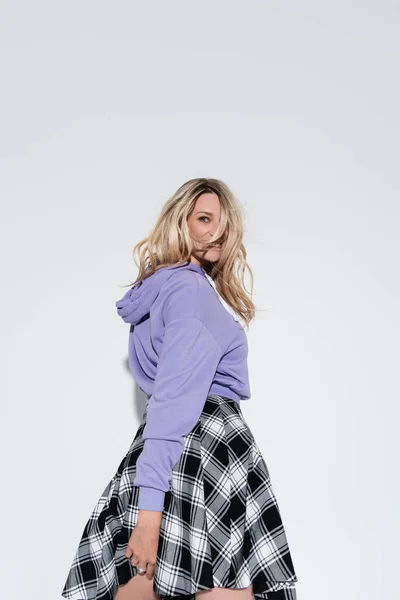 Cheerful blonde woman in trendy purple sweatshirt and plaid skirt posing on grey — Stockfoto